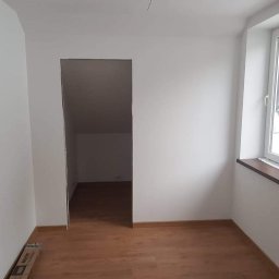 SZWAGRO-BUD - Wnętrze pokoju po malowaniu na biało, widoczne drewniane panele podłogowe, okno z roletą oraz wejście do drugiego pomieszczenia; kartony z materiałami budowlanymi leżą przy ścianie.