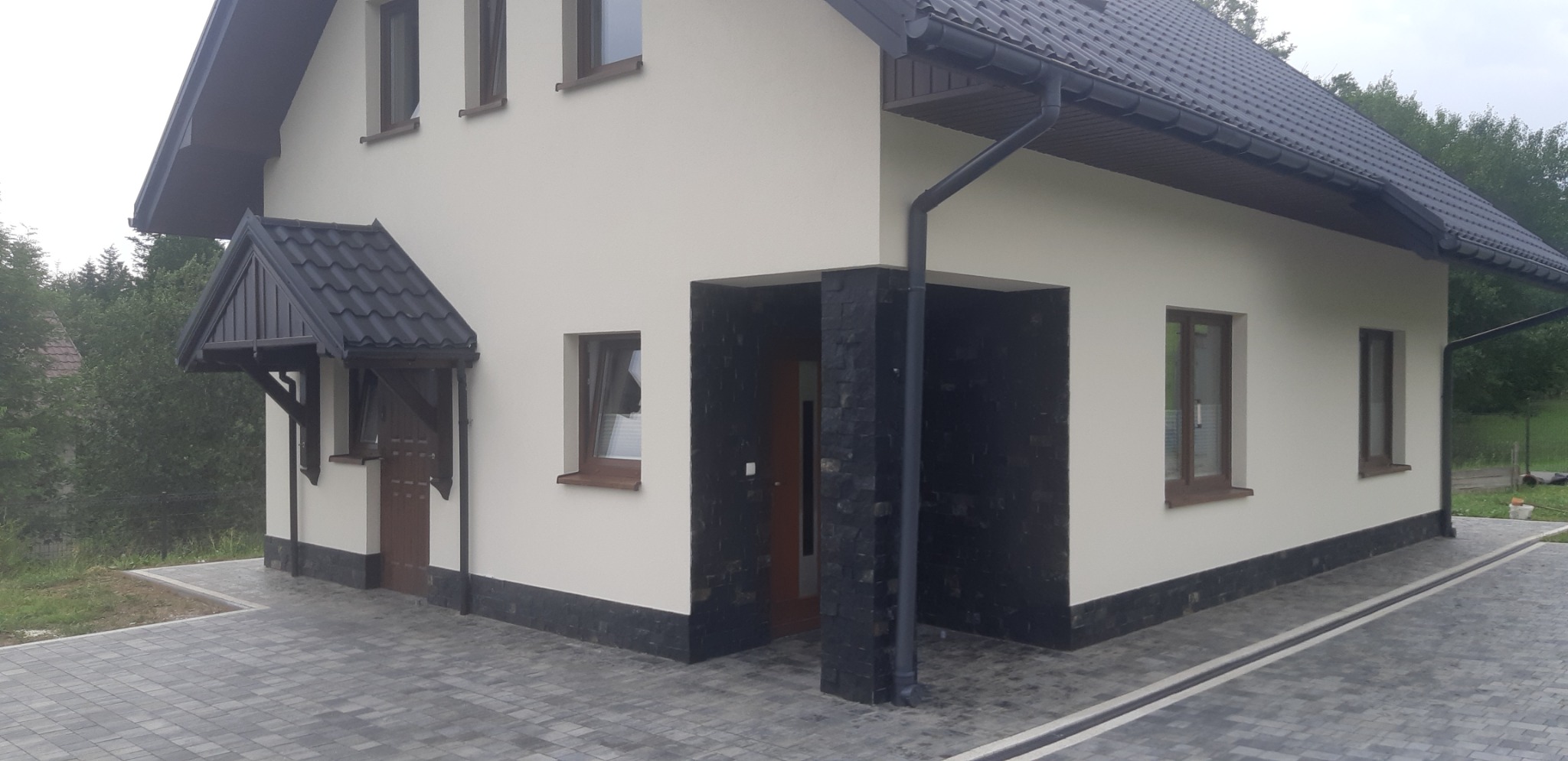 Elewacja domu jednorodzinnego z beżową ścianą, ciemnobrązową stolarką okienną i drzwiami, czarnym kamieniem dekoracyjnym przy wejściu i szarym brukowanym podjazdem.