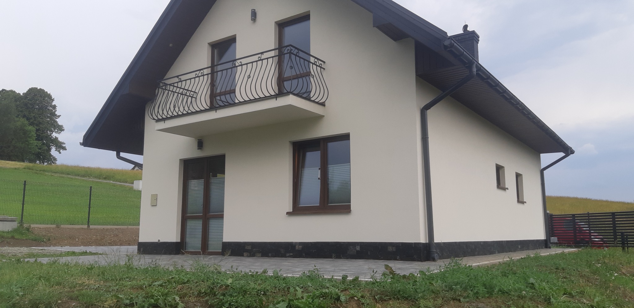 Elewacja domu jednorodzinnego z balkonem i ciemną stolarką okienną, wykończona jasnym tynkiem i ciemną podmurówką, na tle zielonego wzgórza.