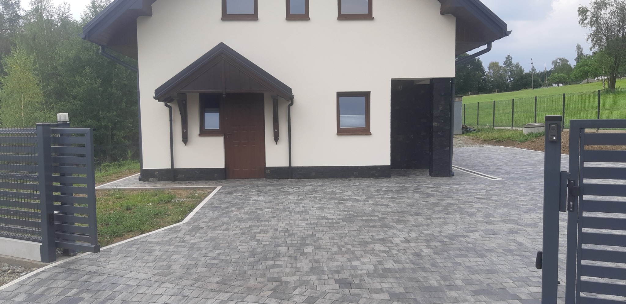 Nowoczesny dom z elewacją w kolorze złamanej bieli, ciemnobrązową stolarką okienną i drzwiami wejściowymi pod zadaszeniem, wykończony kamieniem elewacyjnym przy wjeździe do garażu, z szarą kostką...