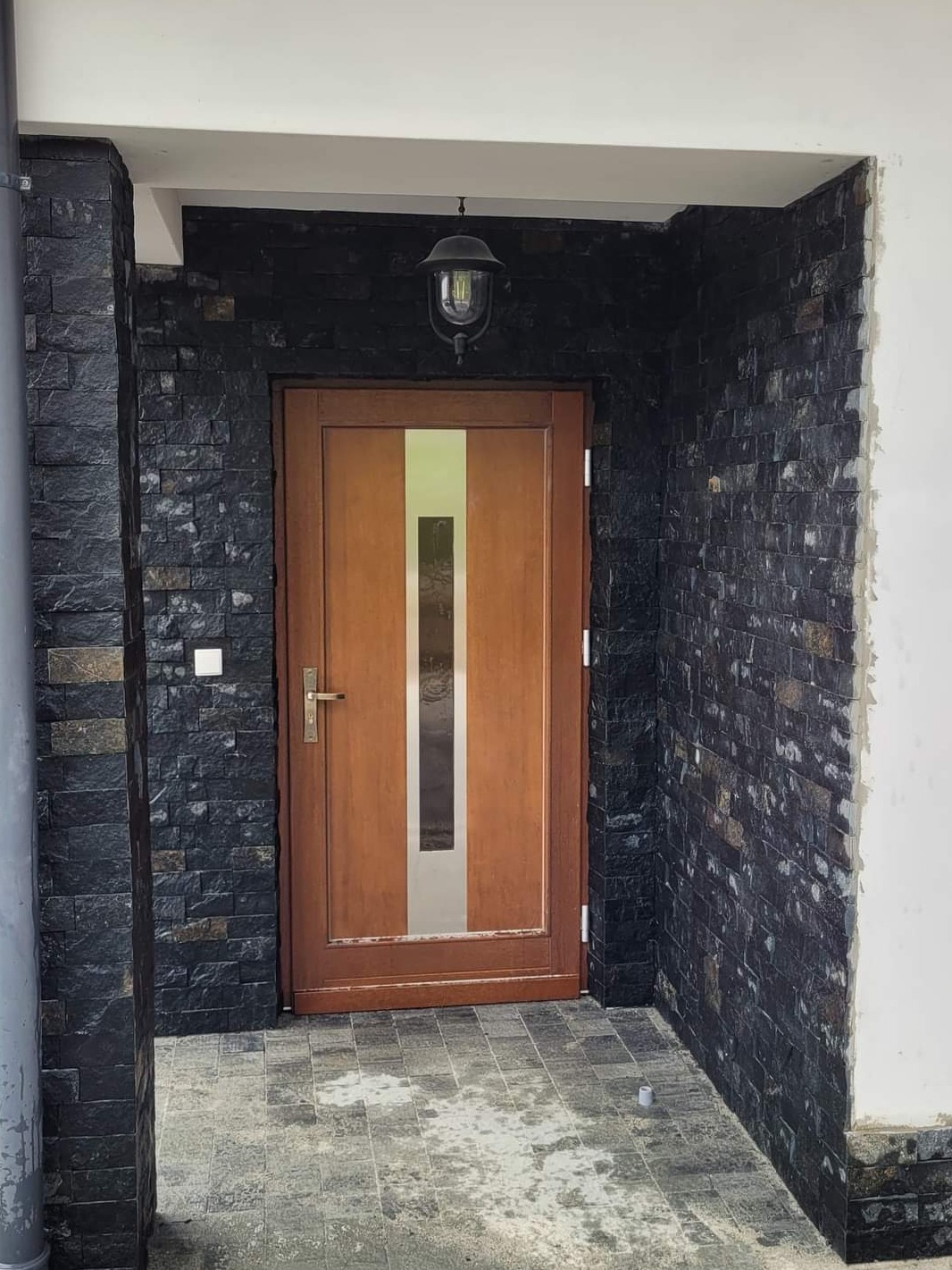 Wejście do domu z drzwiami z pionowym przeszkleniem, obłożone czarnym kamieniem dekoracyjnym, z wiszącą lampą i szarą kostką brukową na podłożu.