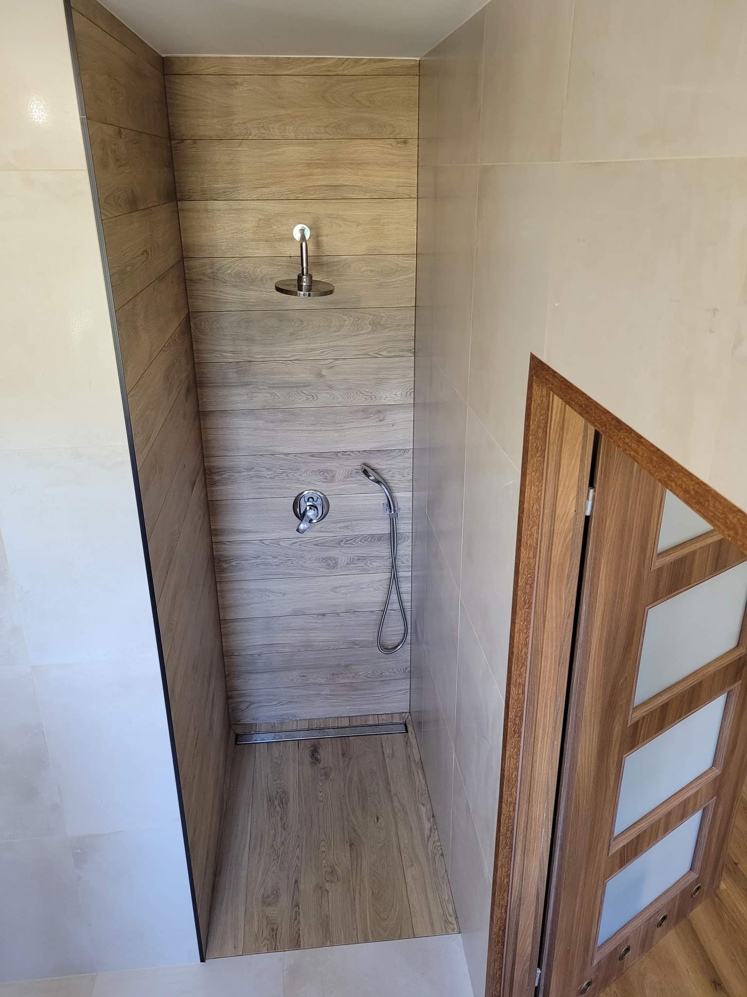 Nowoczesna kabina prysznicowa z panelami imitującymi drewno, chromowaną armaturą i odpływem liniowym, widok z góry na wykończoną łazienkę z elementami drewnopodobnymi.