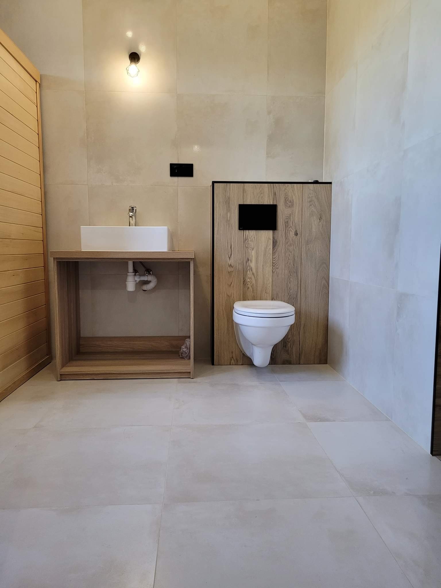 Nowoczesna łazienka z beżowymi płytkami, wiszącą toaletą z drewnianą obudową, prostokątną umywalką na drewnianej szafce i minimalistyczną żarówką