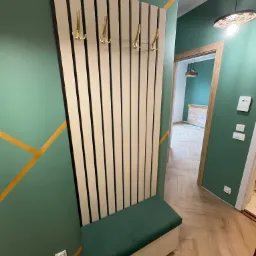 Nowoczesny przedpokój: panel z wieszakami i siedziskiem w odcieniach beżu i zieleni, na tle ściany w geometryczne wzory. Parkiet w jodełkę.