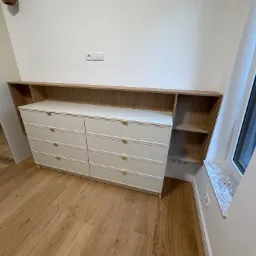 Biała komoda z sześcioma szufladami i drewnianą nadstawką z półkami, ustawiona przy ścianie z kontaktem i oknem z marmurowym parapetem. Jasne drewno na podłodze.