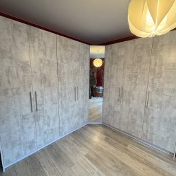 Meble Zbyszek - Szafa wnękowa narożna z lustrem, jasne fronty z delikatnym wzorem, drewniana podłoga, widok z dołu, ciepłe oświetlenie, odbicie osoby w lustrze.