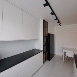 Nowoczesna kuchnia z białymi, lakierowanymi frontami i czarnym blatem. Oświetlenie szynowe podkreśla minimalistyczny design. Widoczny fragment stołu z krzesłami.