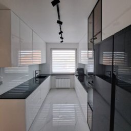Nowoczesna, wąska kuchnia z białymi szafkami i czarnym blatem, oświetlona reflektorami. Okno z żaluzjami w tle. Błyszcząca podłoga odbija światło.