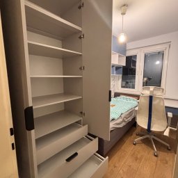 Meble Zbyszek - Otwarta, biała szafa z półkami i szufladami w pokoju, widok z bliska na wnętrze szafy, w tle łóżko i krzesło biurowe, ciepłe oświetlenie.