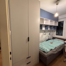 Meble Zbyszek - Jasnoszara szafa z szufladami w pokoju dziecięcym, zintegrowana z zabudową nad łóżkiem i półkami. Na ścianie fototapeta z motywem piłkarskim.