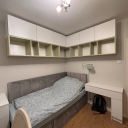 Meble Zbyszek - Białe szafki wiszące z otwartymi półkami nad szarym łóżkiem i biurkiem w pokoju. Oświetlenie sufitowe i lampka biurkowa. Plecak pod biurkiem.