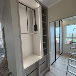 Meble Zbyszek - Wnętrze szafy wnękowej z oświetleniem LED, widoczne półki, szuflady i drążek na ubrania. W tle okno i drabina, sugerujące trwający montaż.