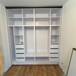 Meble Zbyszek - Biała, otwarta szafa wnękowa z szufladami i półkami, widok z przodu, na podłodze folia ochronna. Widoczne niedoskonałości wykończenia ścian.