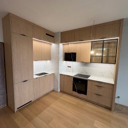 Meble Zbyszek - Jasna kuchnia na wymiar z drewnianymi frontami i marmurowym blatem. Oświetlenie LED pod szafkami. Widok na płytę indukcyjną i piekarnik w zabudowie.