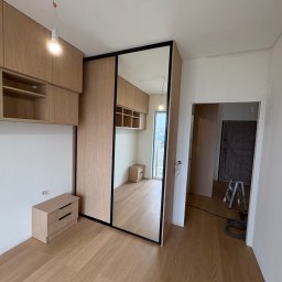 Meble Zbyszek - Jasna szafa wnękowa z lustrem i drewnianymi elementami, widoczna z perspektywy wnętrza. Uzupełniona o szafki wiszące i komodę. W tle widoczne schody i zakryte drzwi.