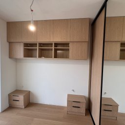 Meble Zbyszek - Jasna szafa wnękowa z lustrzanymi drzwiami przesuwnymi i szafki wiszące nad szafkami nocnymi w pokoju z drewnianą podłogą i oknem balkonowym.