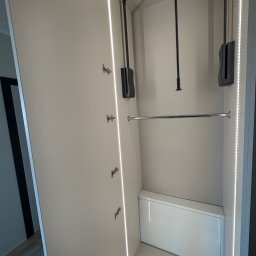 Meble Zbyszek - Otwarta szafa wnękowa z oświetleniem LED po bokach, drążkiem na ubrania i pantografem. Widoczne zawiasy i półka. Minimalistyczny design.