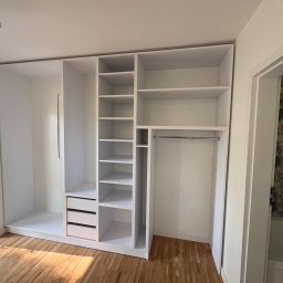 Meble Zbyszek - Biała, modułowa szafa z półkami i szufladami, wbudowana we wnękę pokoju z drewnianą podłogą. Widok na łazienkę z kwiatowym wzorem na ścianie.