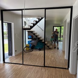 Meble Zbyszek - Nowoczesna szafa wnękowa z czarnymi ramami i lustrzanymi drzwiami, odbijająca wnętrze domu z drewnianymi schodami i podłogą. Widoczny instalator robiący zdjęcie.