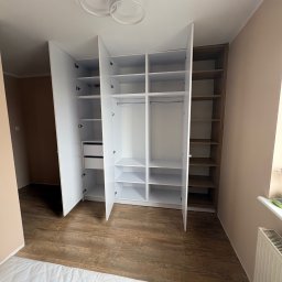 Meble Zbyszek - Jasna szafa wnękowa z otwartymi drzwiami, ukazująca półki i szuflady. Widoczny fragment materaca i grzejnika. Oświetlenie sufitowe w kształcie dwóch pierścieni.