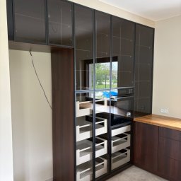 Meble Zbyszek - Nowoczesna zabudowa kuchenna z ciemnego drewna i szklanych frontów, z widocznymi szufladami i piekarnikiem w zabudowie, uzupełniona drewnianym blatem.