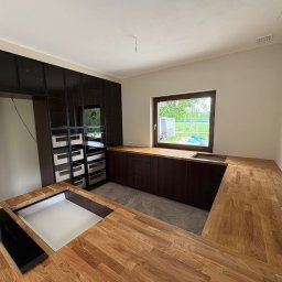 Meble Zbyszek - Nowoczesna kuchnia z drewnianym blatem i czarnymi szafkami, widok na okno z zielenią. Minimalistyczny design, jasne wnętrze, wbudowana płyta indukcyjna i zlew.
