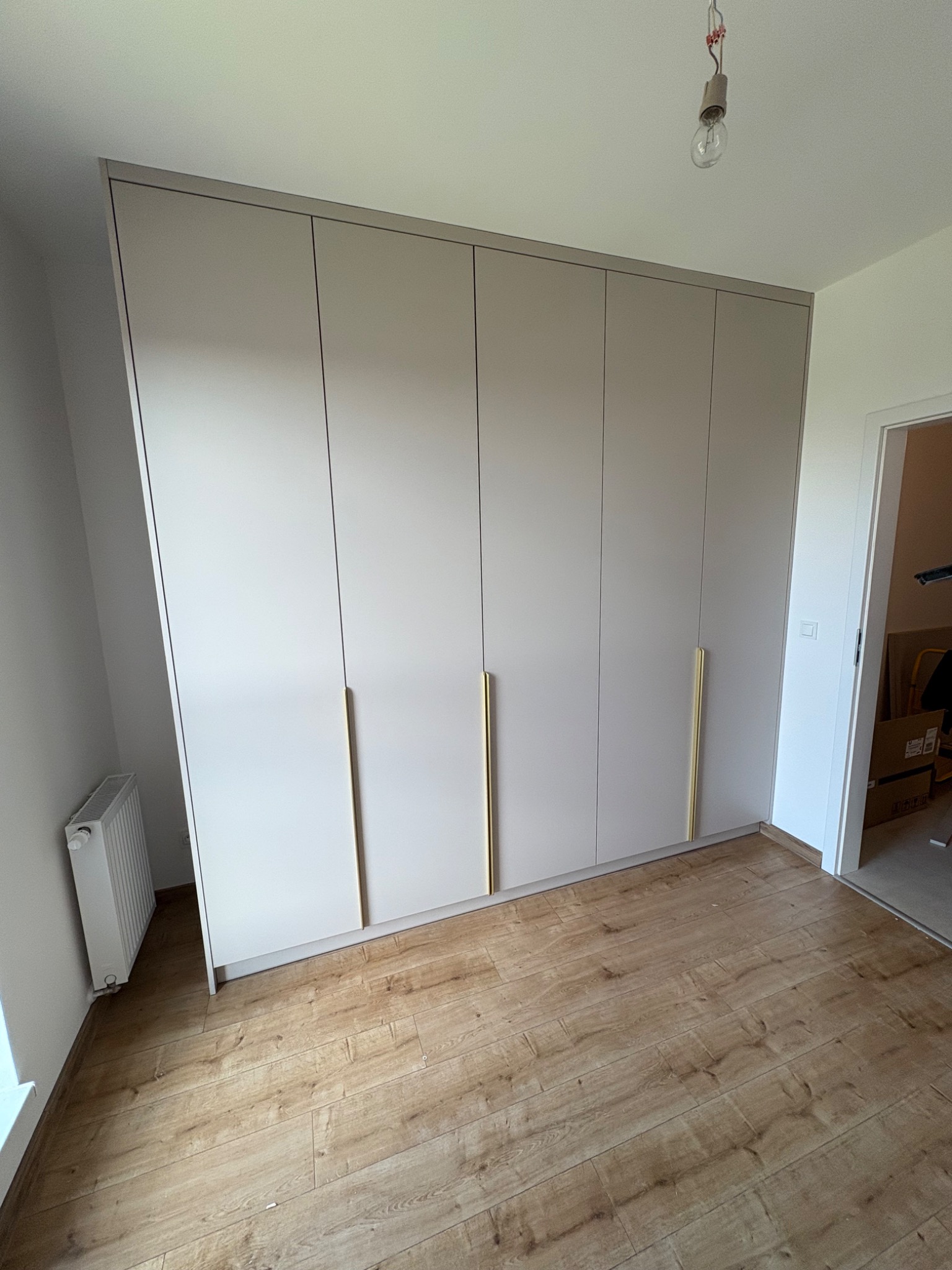 Beżowa szafa wnękowa z długimi, złotymi uchwytami, na drewnianej podłodze. Widoczny grzejnik i surowa żarówka.