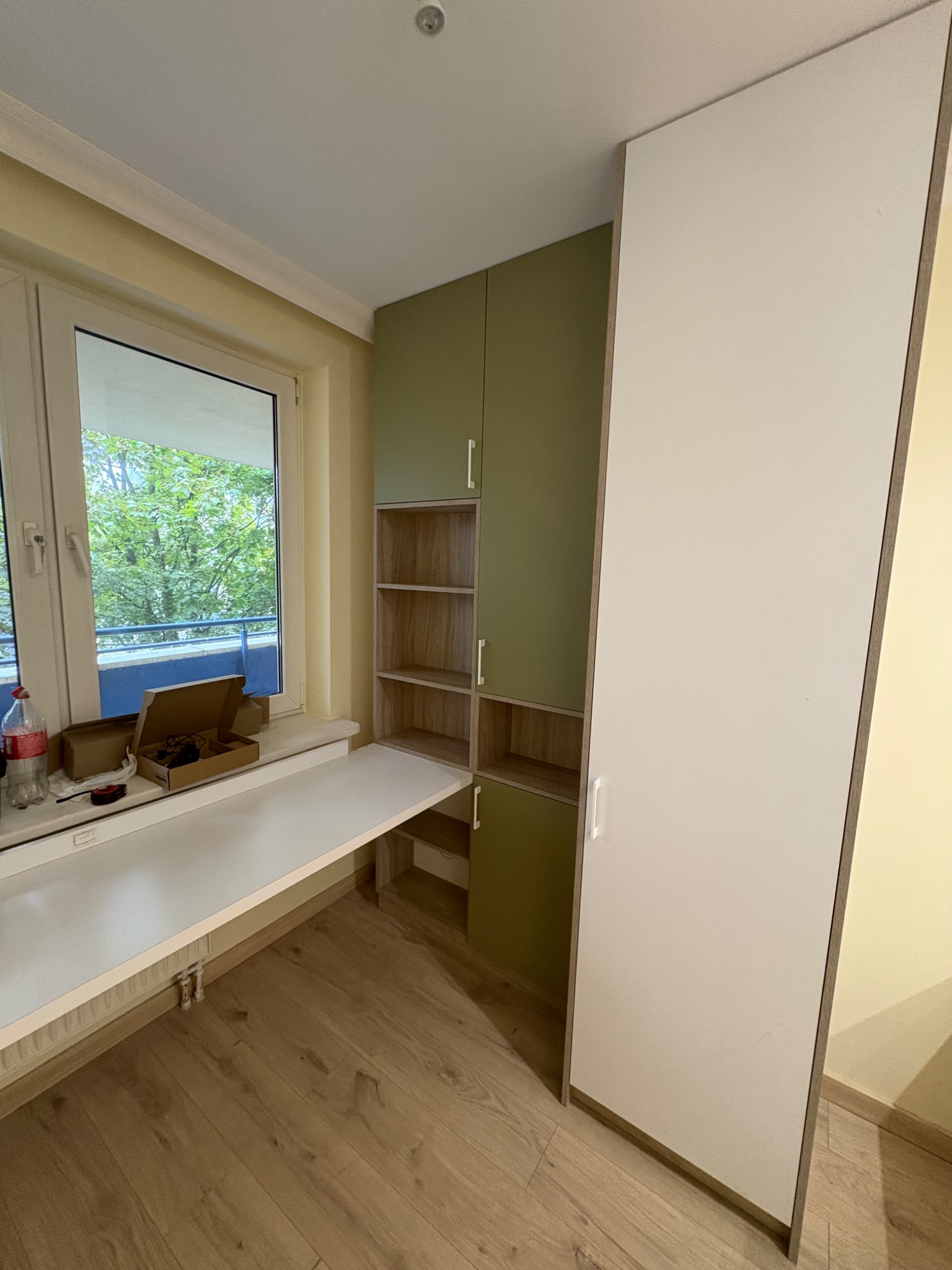 Zabudowa na wymiar z szafą, biurkiem pod oknem i półkami w odcieniach bieli, zieleni i drewna. Widok na zieleń za oknem. Na blacie leżą narzędzia i pudełka.