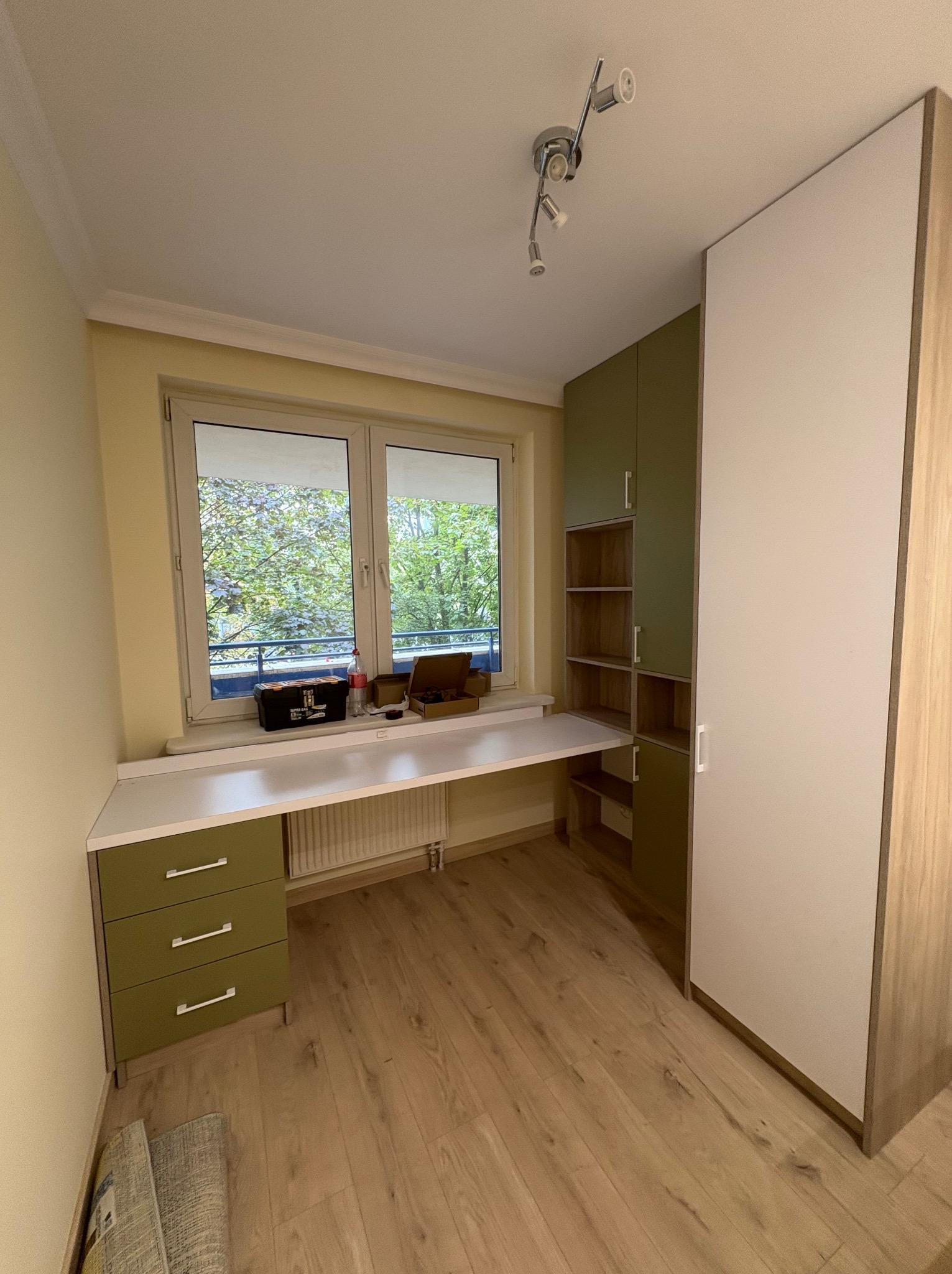 Nowoczesna szafa z biurkiem na wymiar w pokoju, z zielonymi akcentami i widokiem na zieleń za oknem. Minimalistyczny design, jasne drewno i białe fronty.