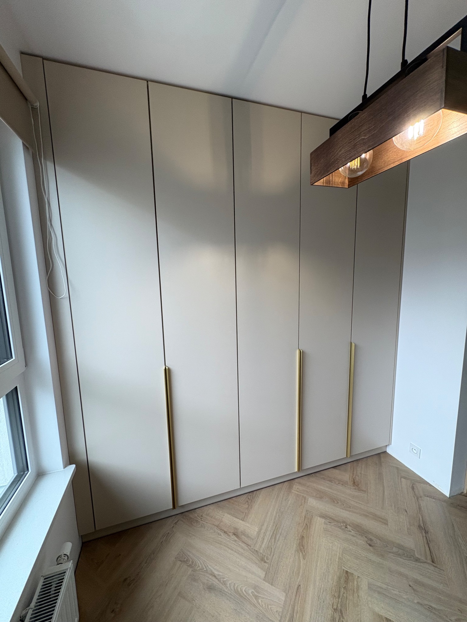 Jasna szafa wnękowa z złotymi uchwytami, podłoga w jodełkę. U góry widoczna lampa z drewnianymi elementami. Minimalistyczny design.