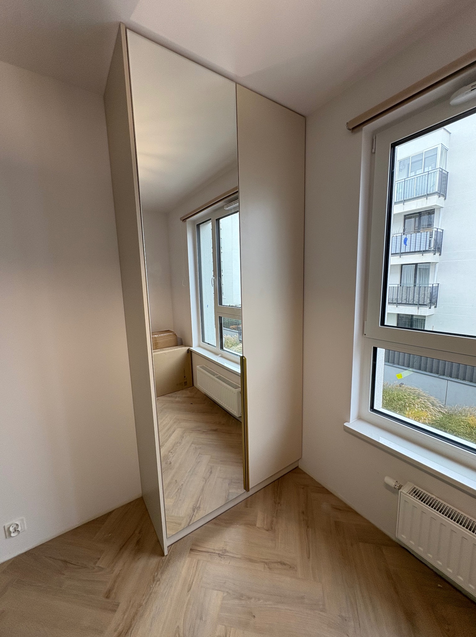 Jasna szafa wnękowa z lustrem, w rogu pokoju z parkietem w jodełkę. Widok na okno i budynek za nim. Minimalistyczny design, neutralna kolorystyka.