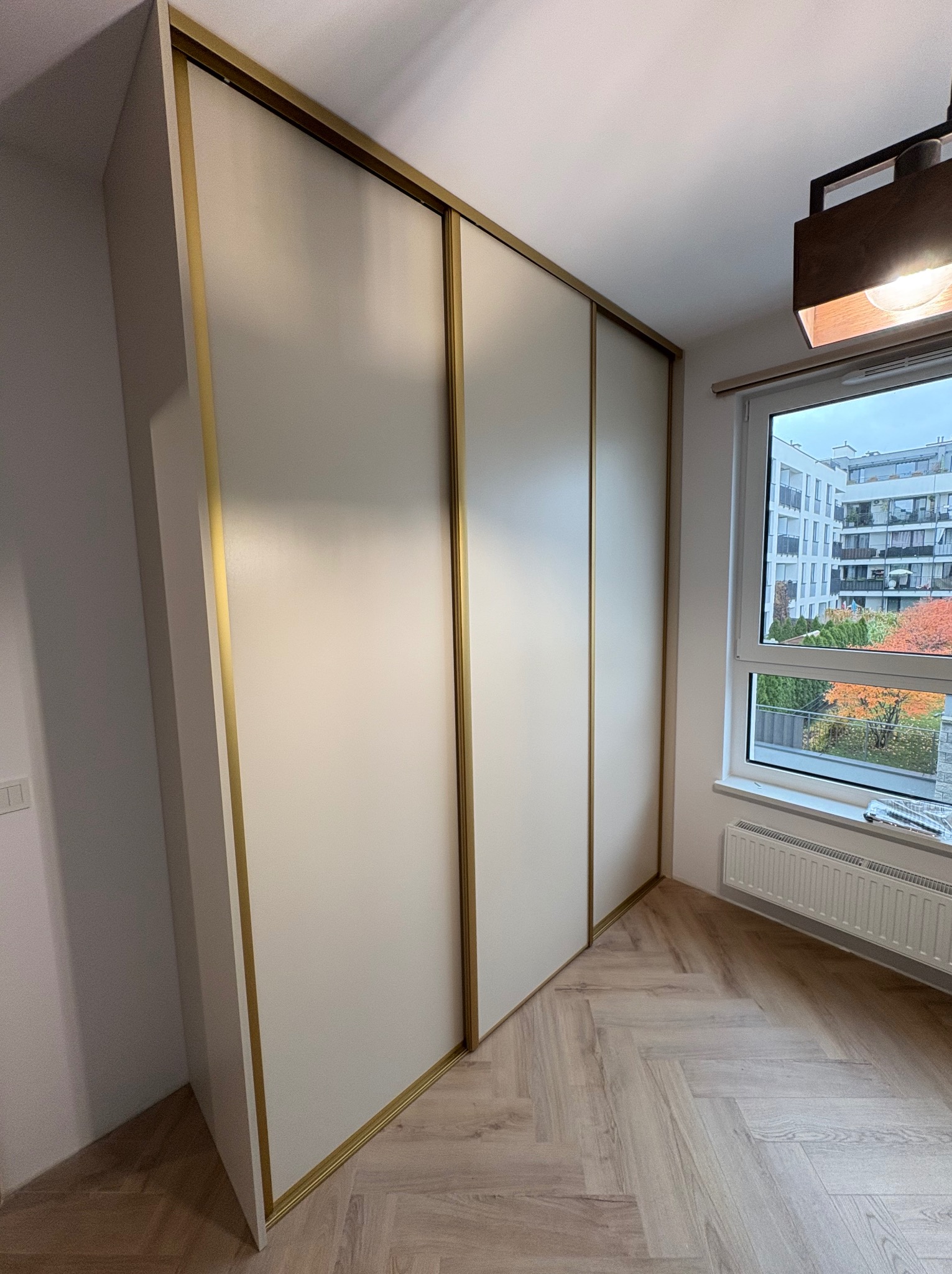 Trzydrzwiowa szafa wnękowa z przesuwnymi drzwiami i złotymi obramowaniami, widok z boku, na podłodze parkiet w jodełkę, w tle okno z widokiem na budynek.