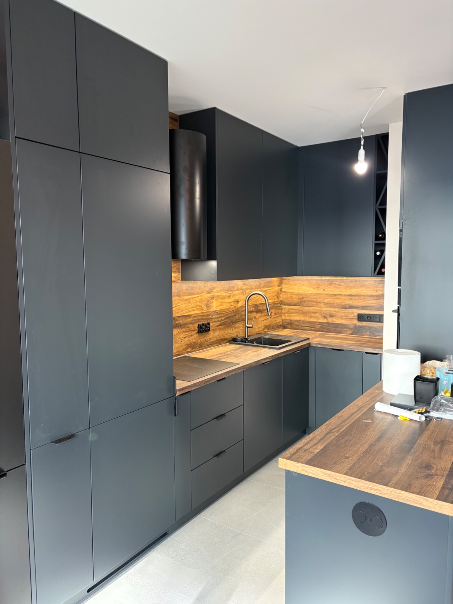 Nowoczesna kuchnia z ciemnymi, matowymi frontami i drewnianym blatem. Na blacie widoczny zlew i płyta, a w tle półka na wino. Minimalistyczny design.