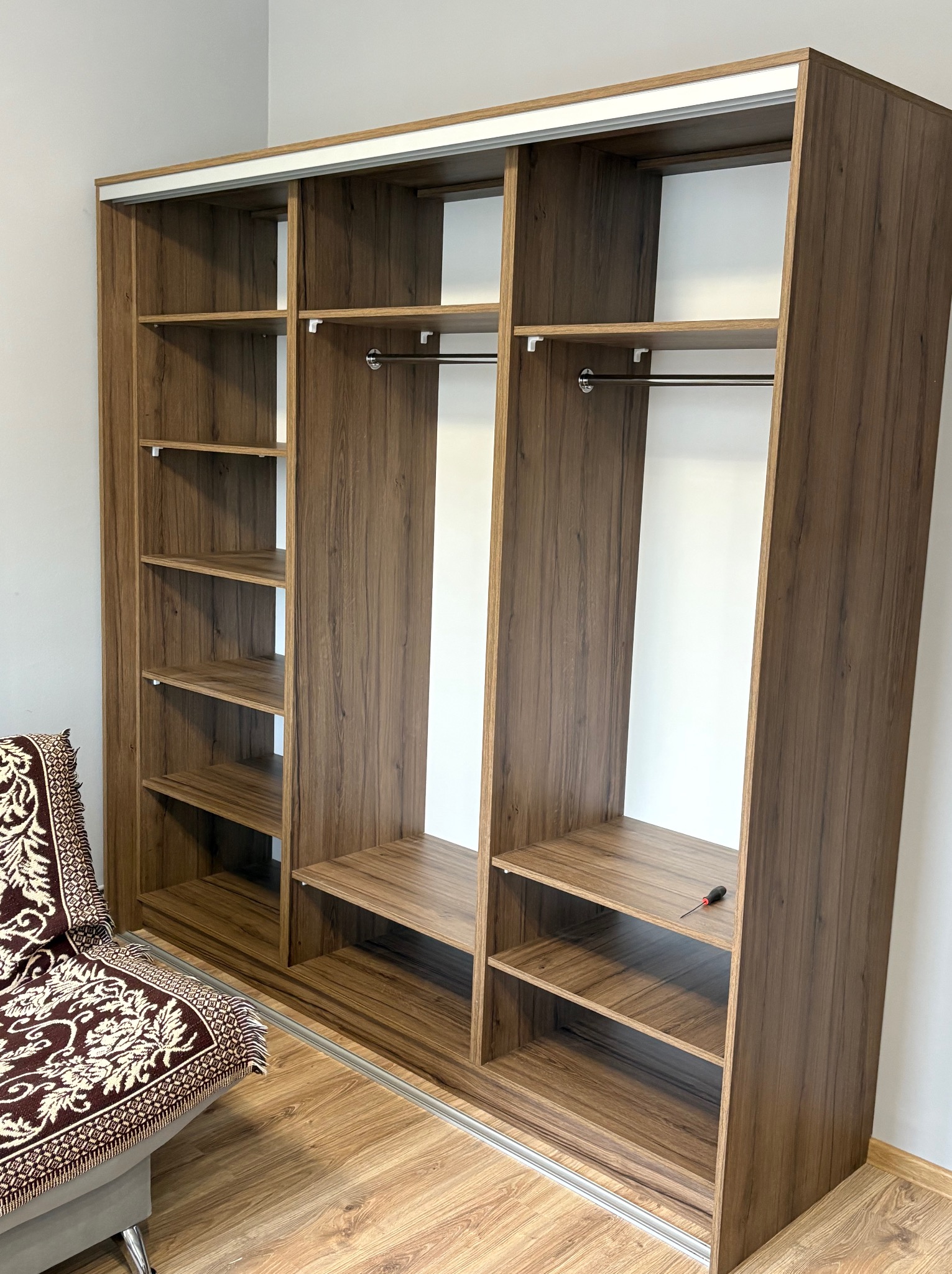 Otwarta szafa przesuwna z półkami i drążkami, wykończona okleiną drewnopodobną. Na półce leży śrubokręt, obok stoi fotel z narzutą w kwiaty.