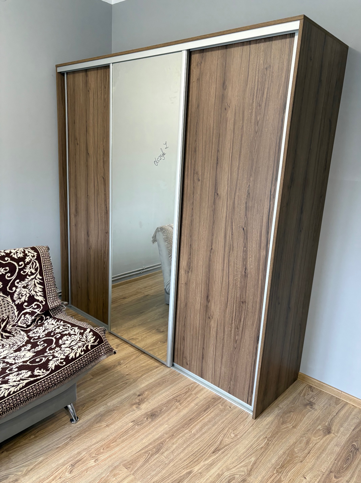 Szafa przesuwna z lustrem i drewnianymi frontami, widoczna z boku w pokoju. Na lustrze widoczny napis. Obok stoi kanapa z wzorzystą narzutą.
