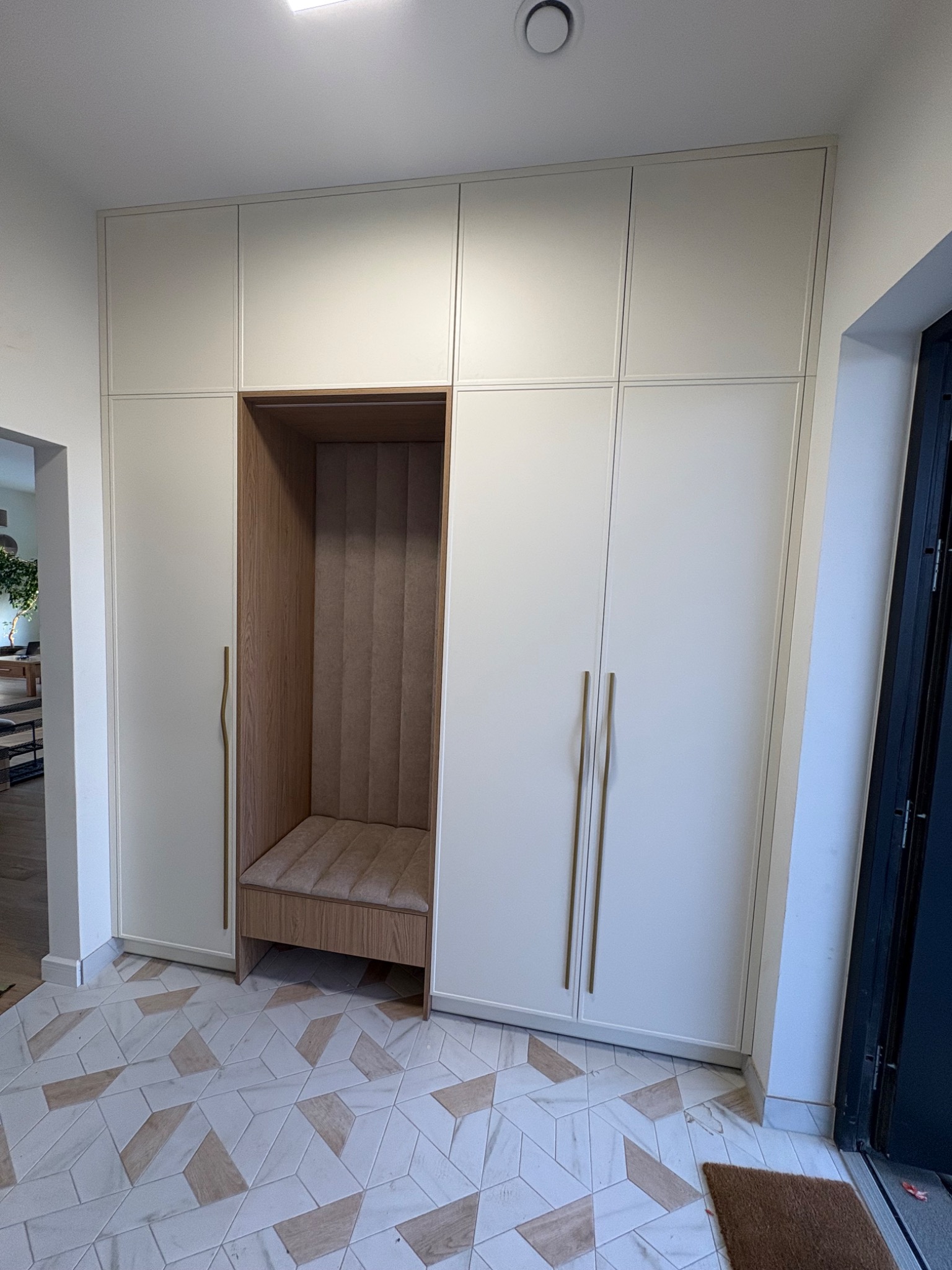 Biała szafa wnękowa z siedziskiem i obiciem w kolorze beżowym. Złote, smukłe uchwyty. Geometryczna posadzka. Nowoczesny design.