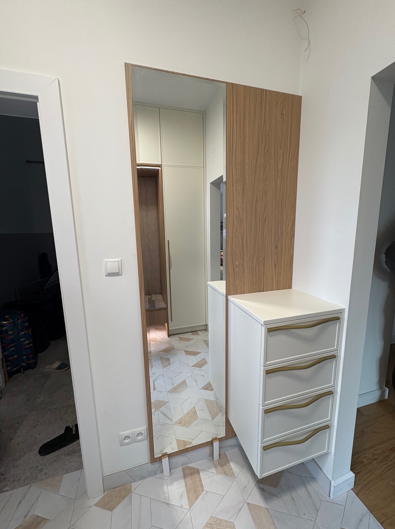 Nowoczesna szafa wnękowa z lustrem i komodą w przedpokoju, wykończona drewnem i białym lakierem, z widocznymi złotymi uchwytami szuflad.