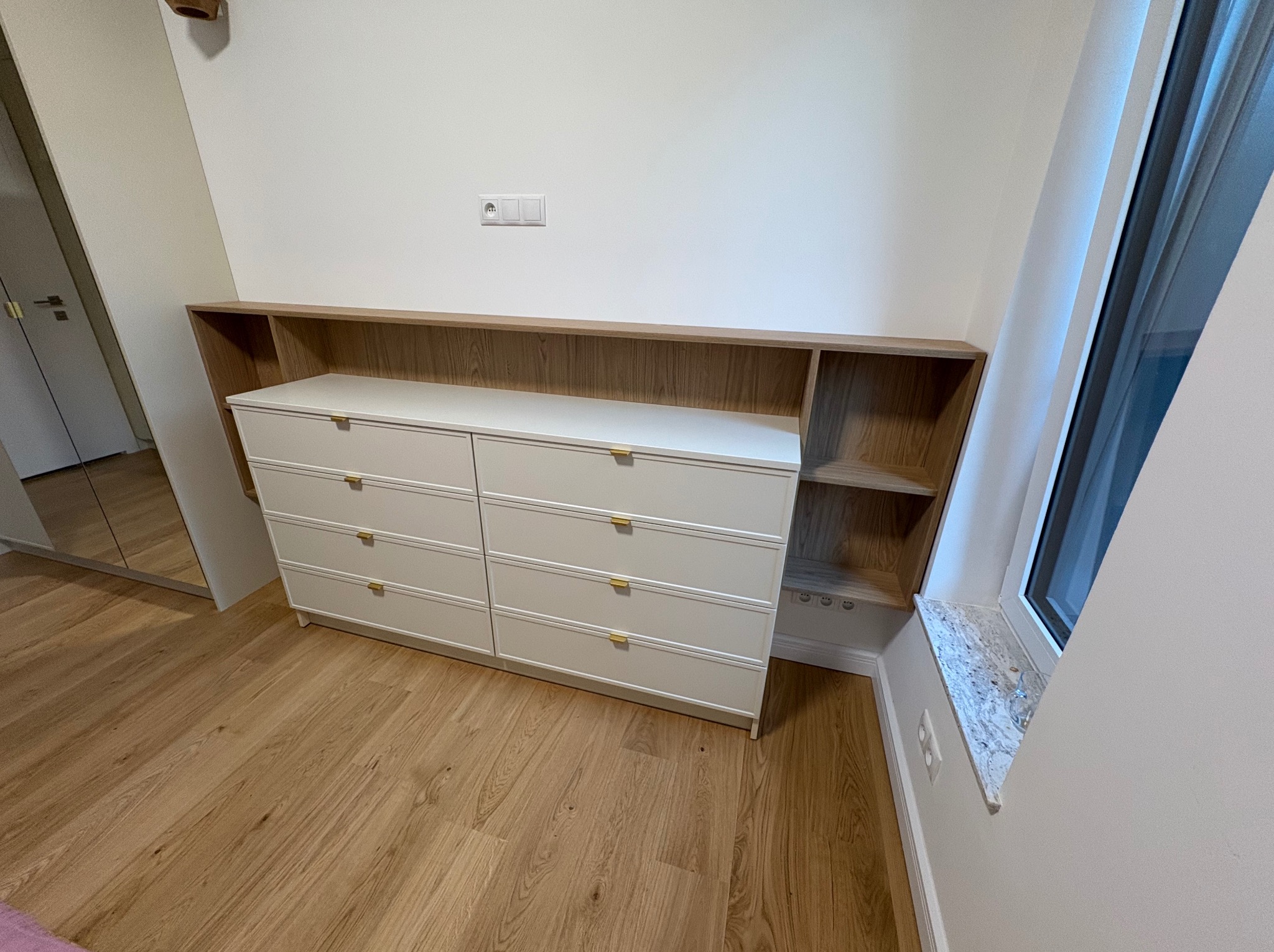 Biała komoda z sześcioma szufladami i drewnianą nadstawką z półkami, ustawiona przy ścianie z kontaktem i oknem z marmurowym parapetem. Jasne drewno na podłodze.