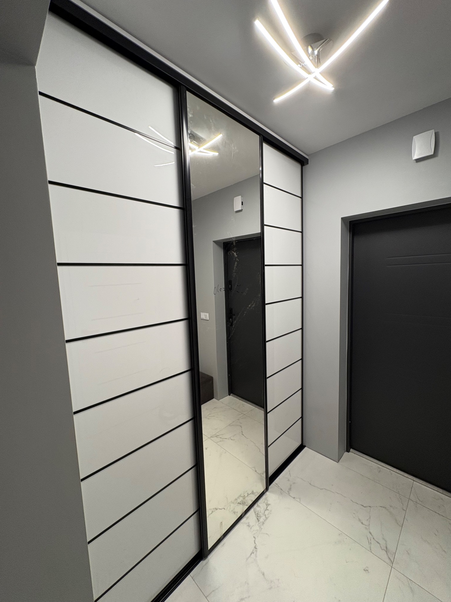 Wnętrze przedpokoju z białą szafą wnękową z przesuwnymi drzwiami z lustrem, czarne akcenty, marmurowa podłoga, nowoczesne oświetlenie sufitowe.