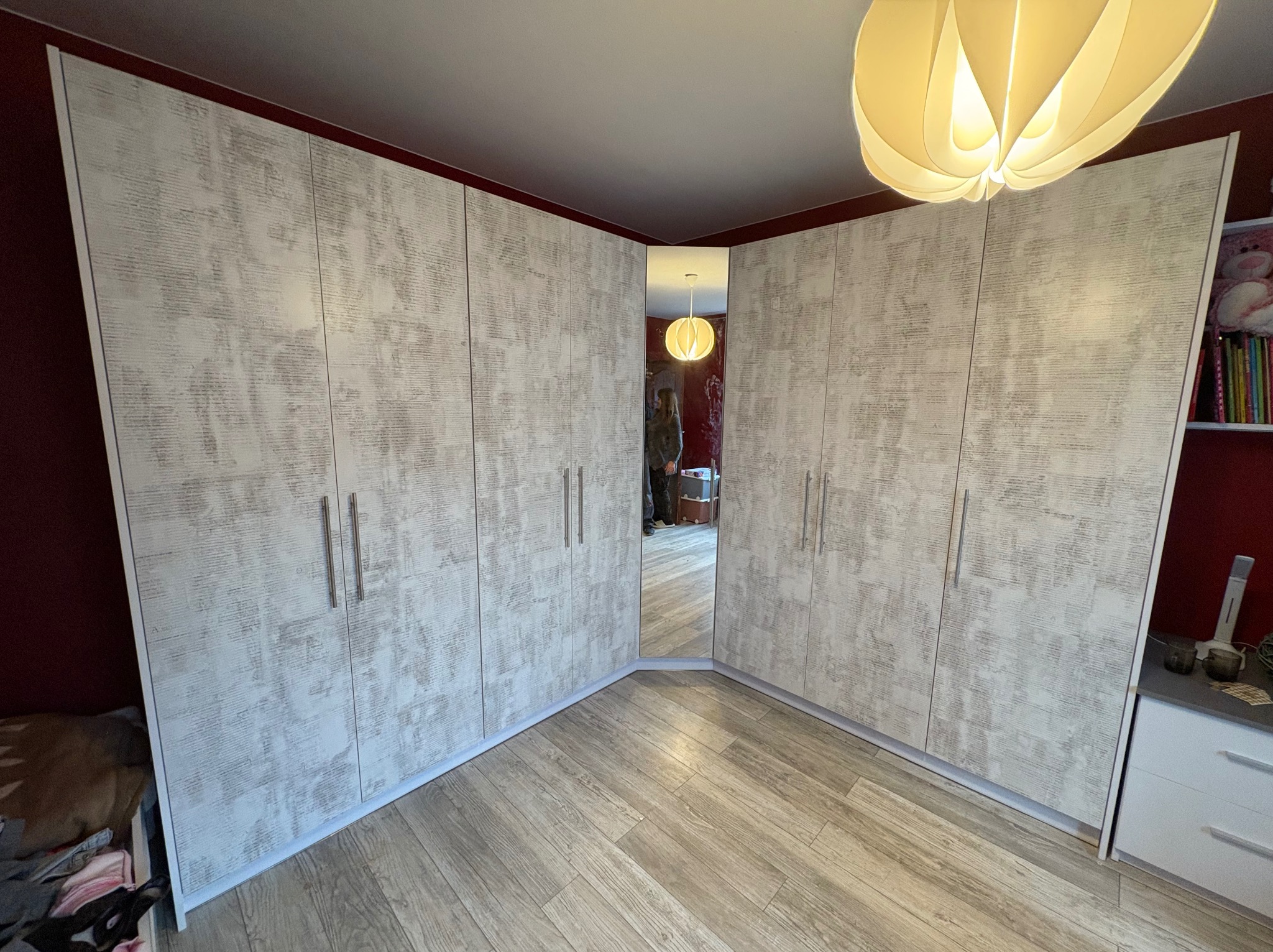 Szafa wnękowa narożna z lustrem, jasne fronty z delikatnym wzorem, drewniana podłoga, widok z dołu, ciepłe oświetlenie, odbicie osoby w lustrze.