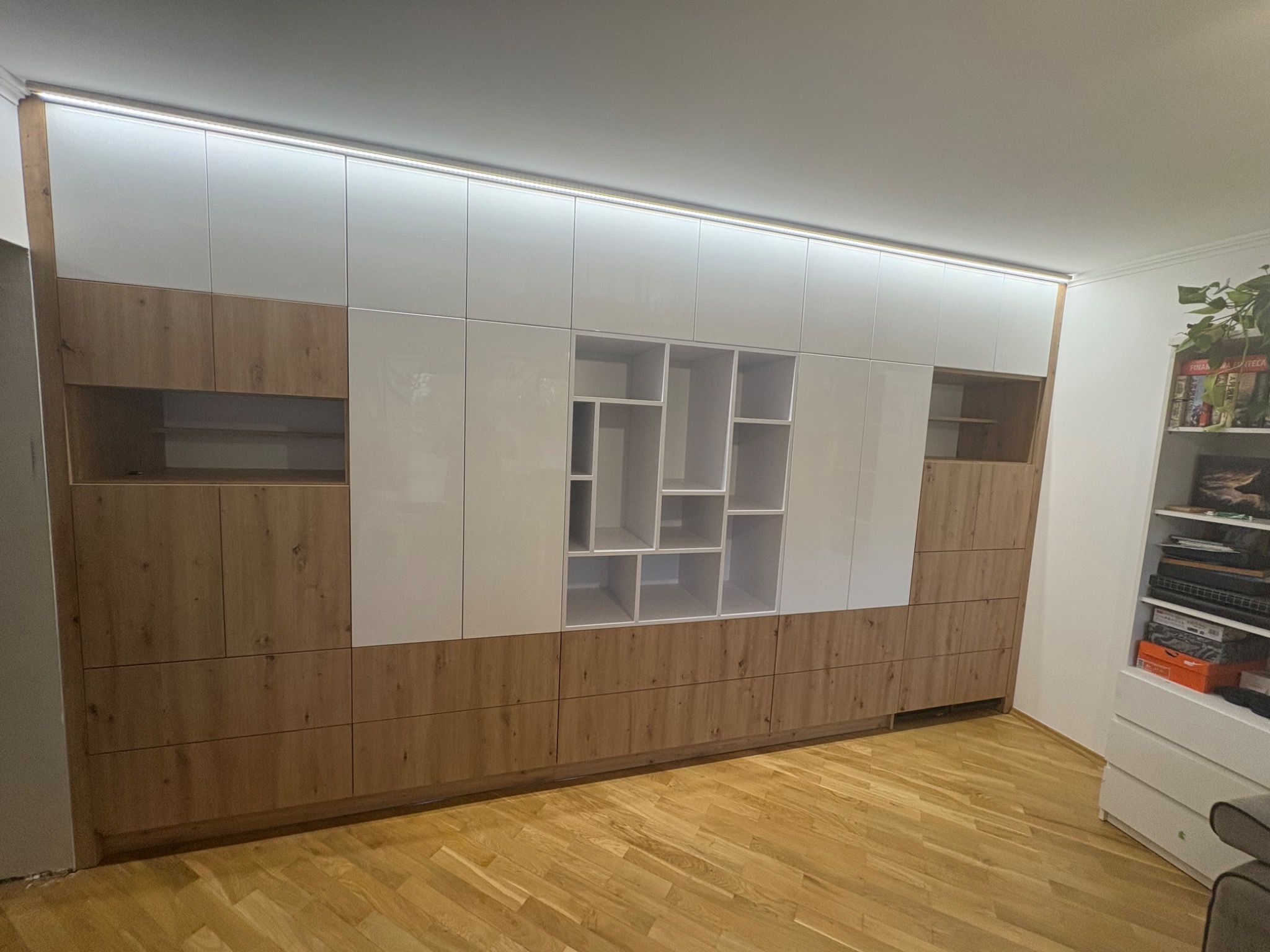 Nowoczesna szafa wnękowa z białymi, lakierowanymi frontami i drewnopodobnymi panelami dolnymi. Centralna część z otwartymi półkami o nieregularnym układzie.