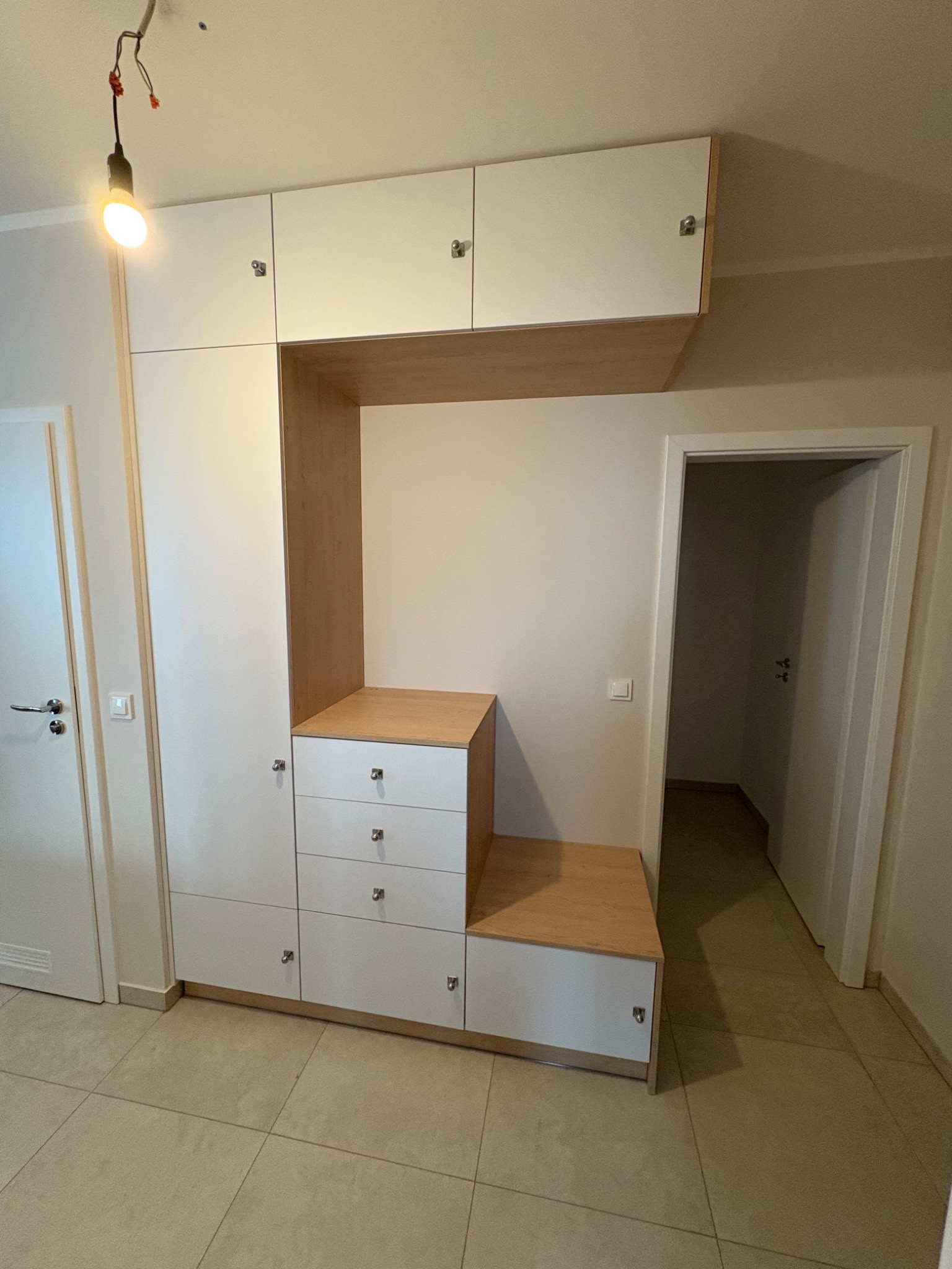 Biała szafa wnękowa z drewnianymi akcentami i szufladami, wkomponowana w przedpokój z beżową podłogą i widocznym wejściem do innego pomieszczenia.