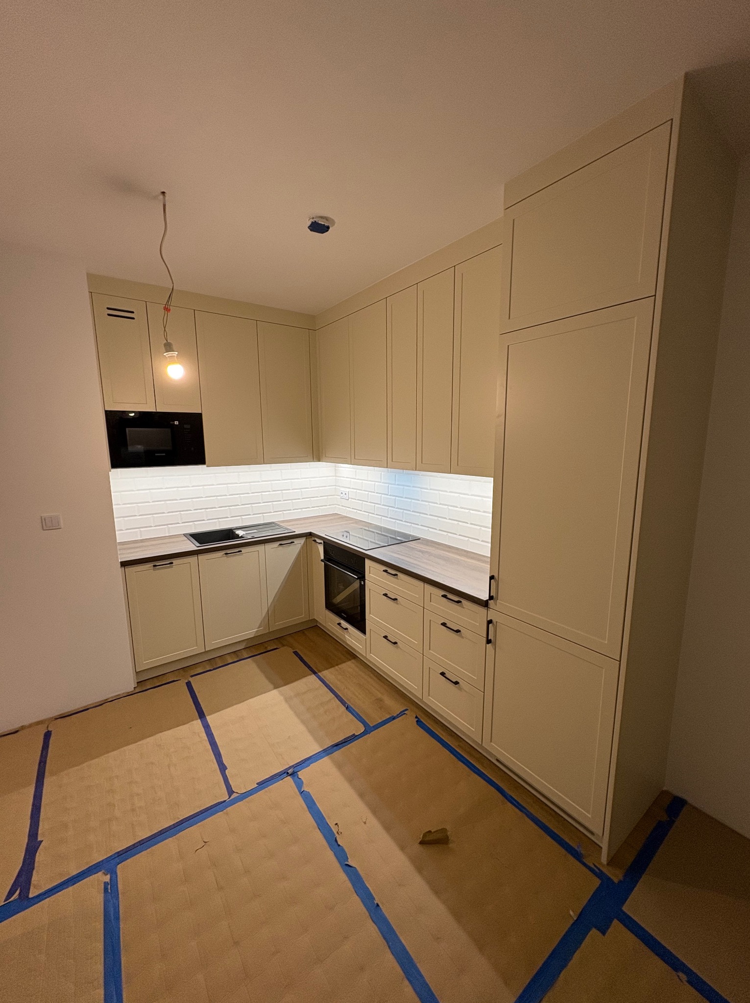 Jasna kuchnia na wymiar z białymi płytkami metra i drewnianym blatem. Podłoga zabezpieczona kartonem i taśmą malarską. Widoczna lampa bez klosza.
