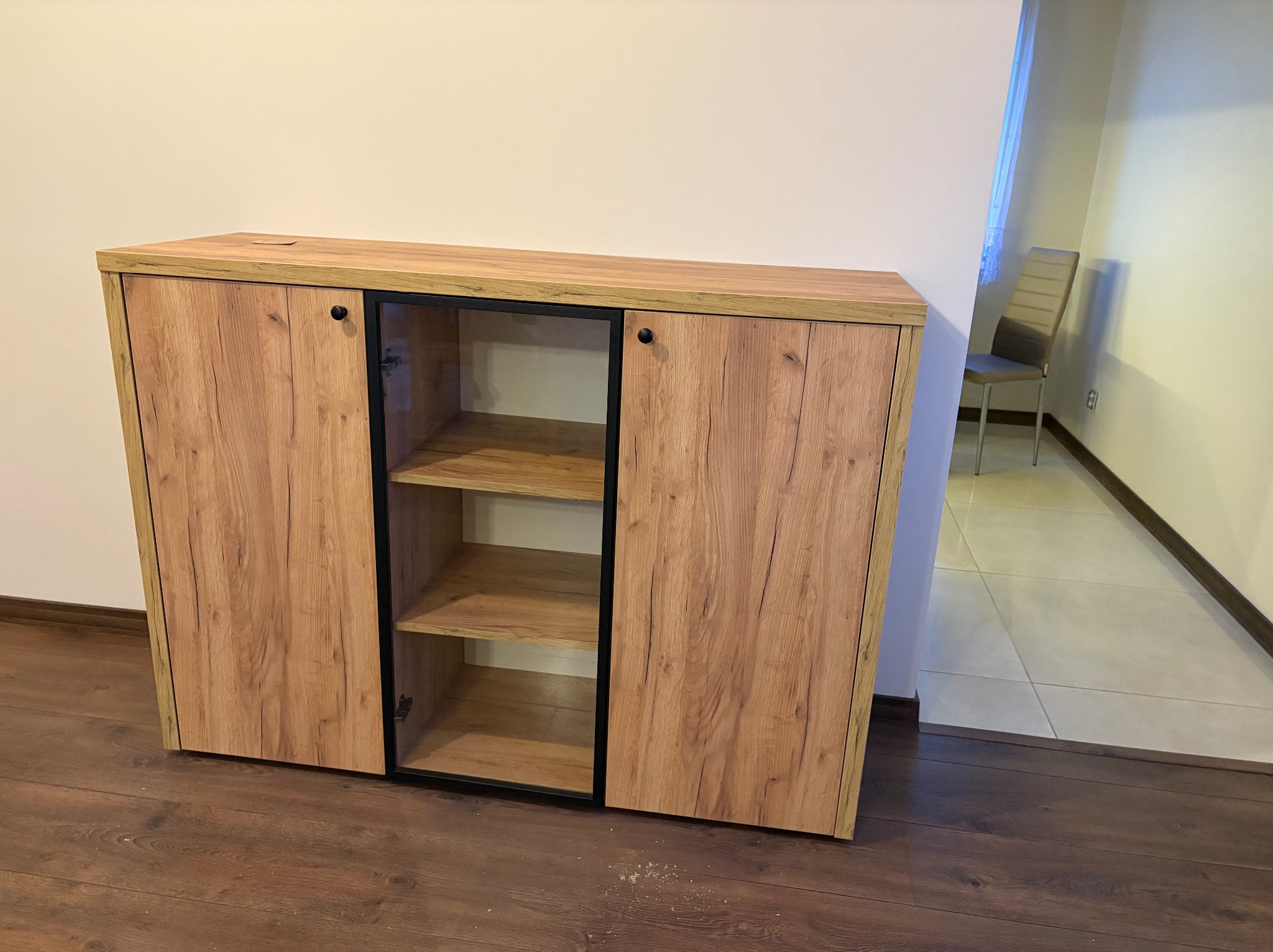 Drewniana komoda z przesuwnymi drzwiami i półkami w środku, wykonana na wymiar w Radomsku. Widoczne usłojenie drewna i czarne obramowanie.