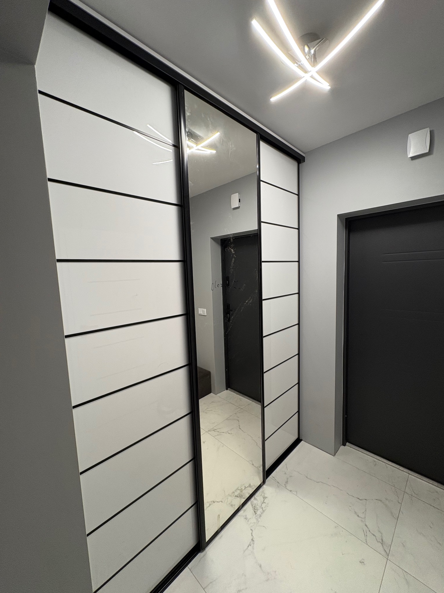Nowoczesna szafa wnękowa z lustrem i białymi panelami, czarne akcenty, marmurowa podłoga, oświetlenie sufitowe w kształcie gwiazdy, szare ściany i czarne drzwi.
