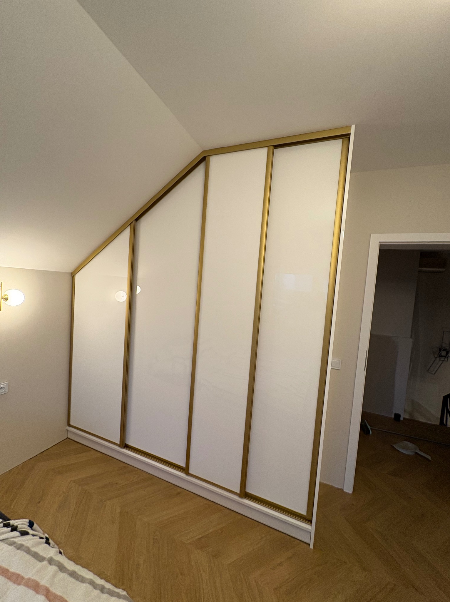 Biała szafa wnękowa z przesuwnymi drzwiami i złotymi ramami, dopasowana do skosu dachu, na tle parkietu ułożonego w jodełkę.