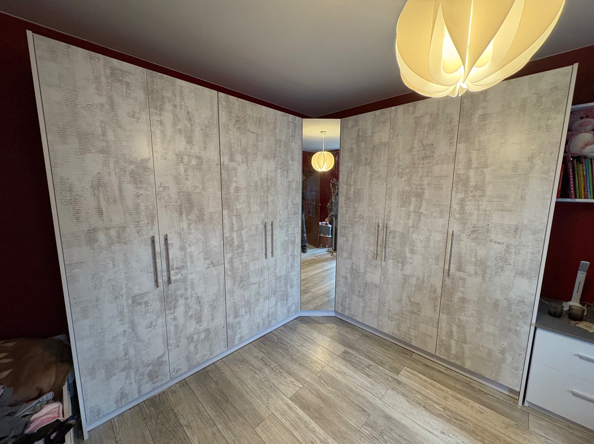 Jasna szafa narożna z lustrem wkomponowana w przestrzeń pokoju, widok z perspektywy podłogi, oświetlona lampą wiszącą. Fronty szafy z delikatnym wzorem.