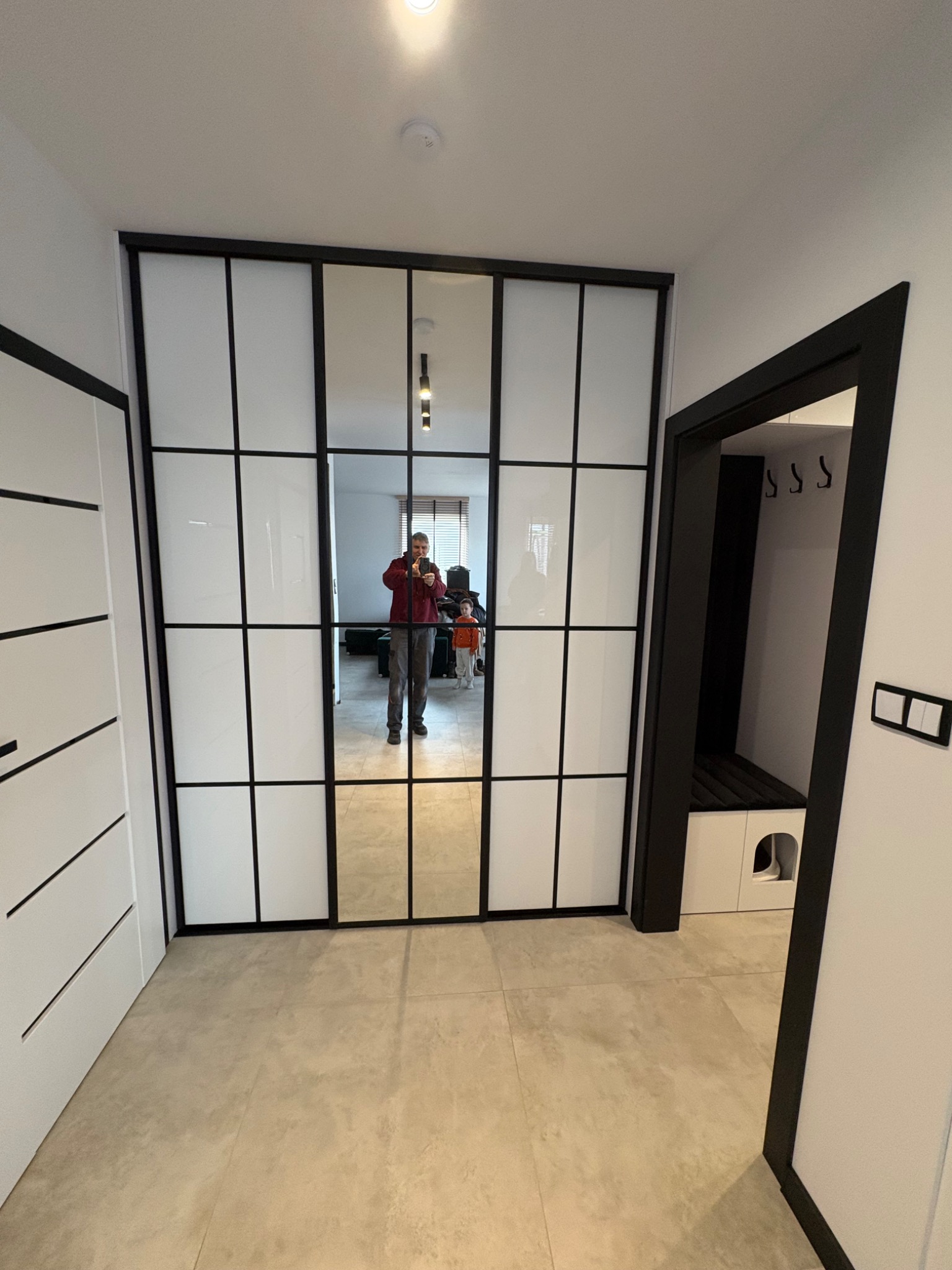 Nowoczesna szafa wnękowa z lustrzanymi drzwiami w czarnej ramie, odbijająca wnętrze mieszkania i osobę robiącą zdjęcie. Minimalistyczny design, jasne kolory.