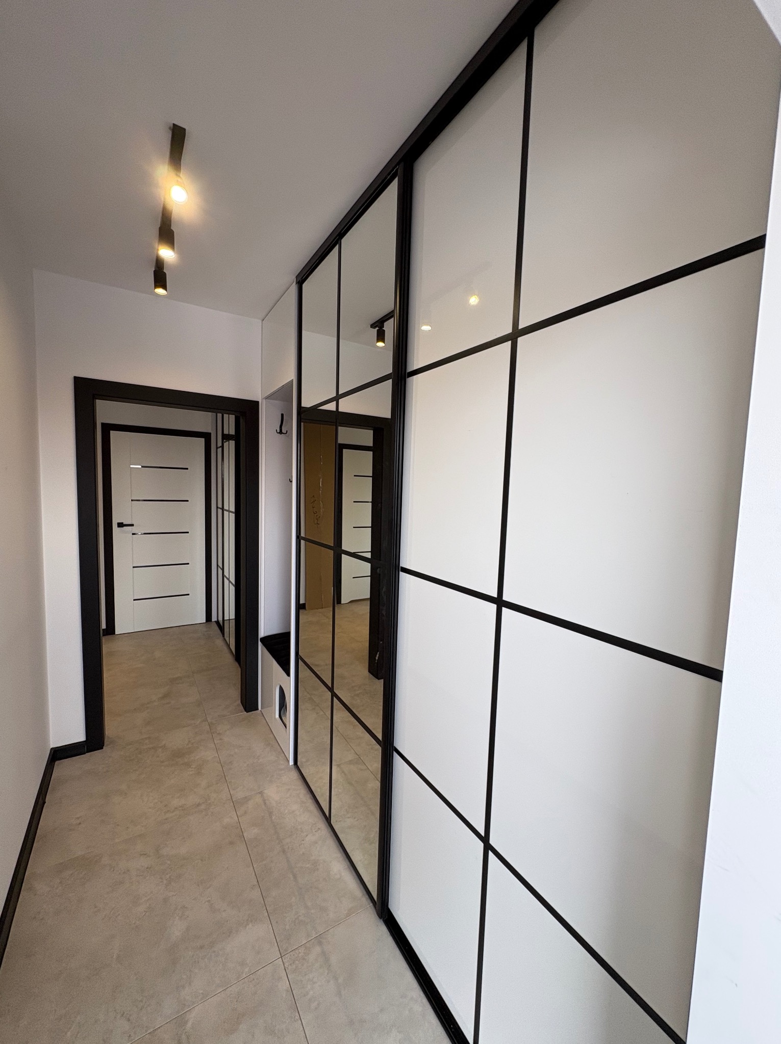 Nowoczesna szafa wnękowa z przesuwnymi drzwiami z lustrami i białymi panelami, czarne ramy, oświetlenie punktowe, widok na przedpokój z drzwiami.