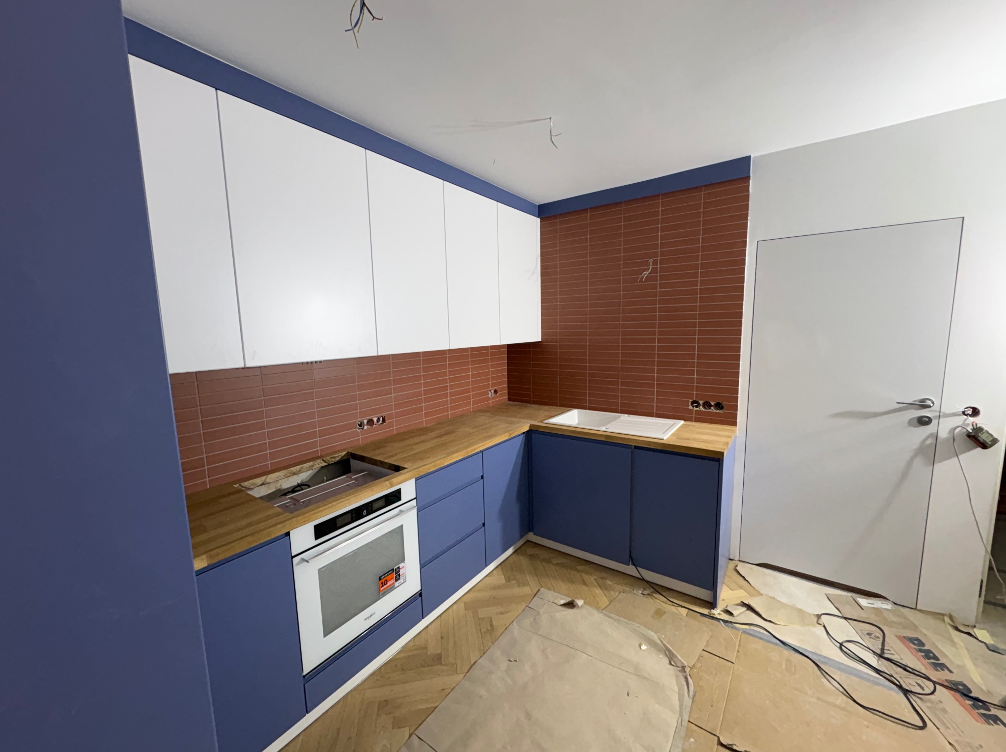 Niebiesko-biała kuchnia z drewnianym blatem i ceglaną ścianą w trakcie montażu. Widoczne zabezpieczenia podłogi i wystające kable.